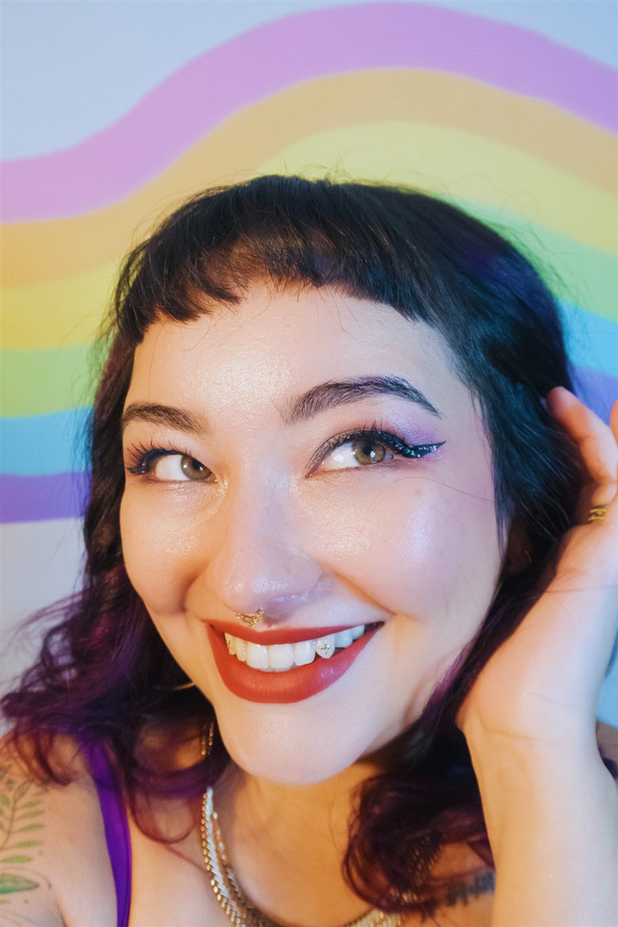 Foto da Bella, fundadora do Ninho: uma mulher sorridente, de cabelo escuro com franja, batom vermelho e piercing dourado no nariz, em frente a uma parede pintada com um arco-íris.